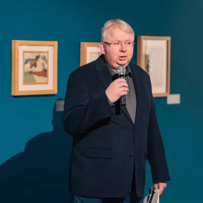 Portret mężczyzny trzymającego  w prawej ręce mikrofon. Mężczyzna ubrany jest w marynarkę, ma na twarzy okulary, w lewej dłoni trzyma teczkę. W tle na ścianie wiszą obrazy.