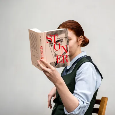 Osoba o rudych włosach siedzi na krześle, trzyma przed twarzą otwartą książkę pt. „Stoner” autorstwa Johna Williamsa. Okładka książki z ilustracją męskiej twarzy niemal idealnie pokrywa się z głową czytelnika, tworząc złudzenie, że narysowana postać jest częścią rzeczywistej osoby.